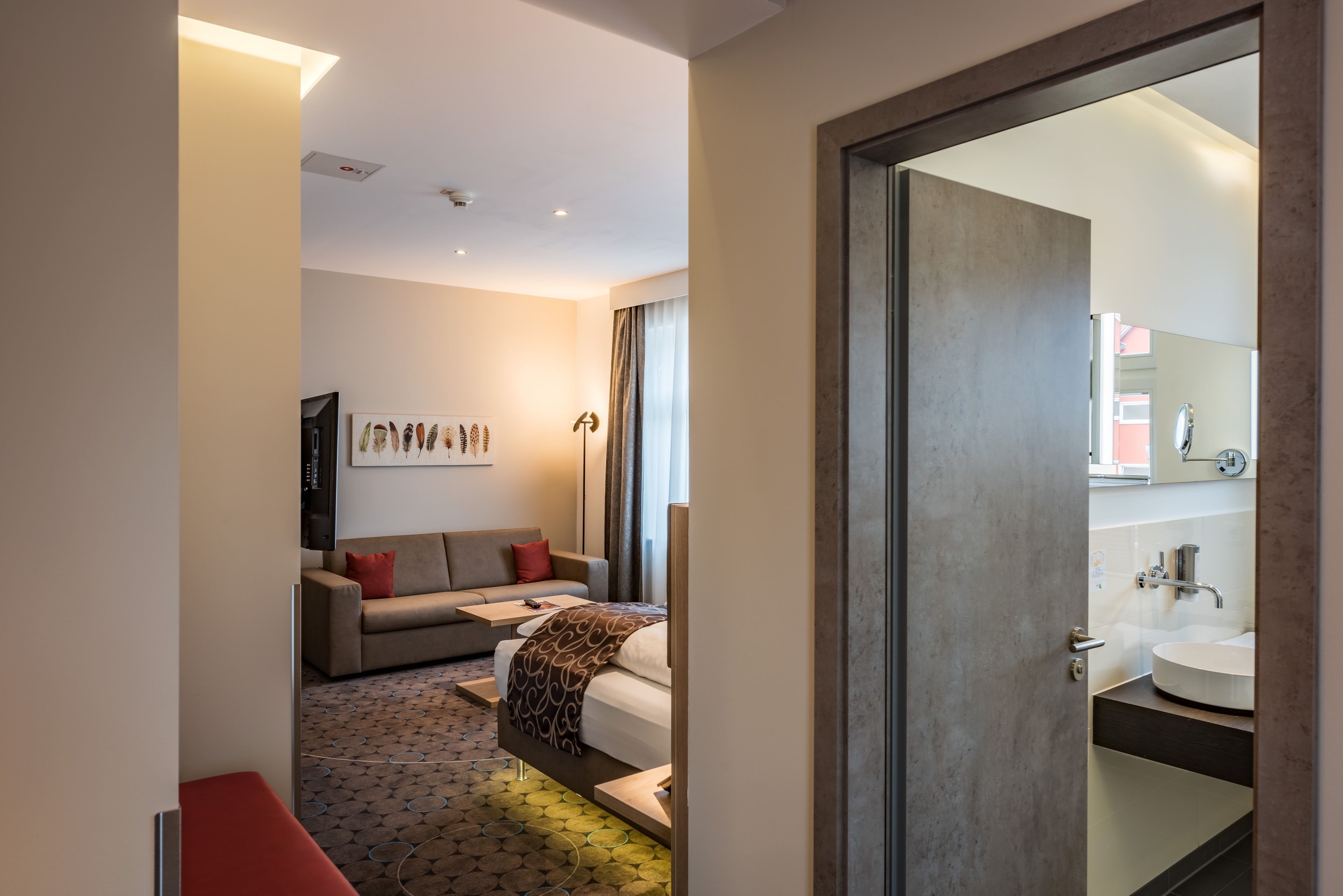 Une chambre d’hôtel moderne, équipée d’un lit, d’un canapé, d’une petite table et d’une salle de bain avec un lavabo et un miroir.
