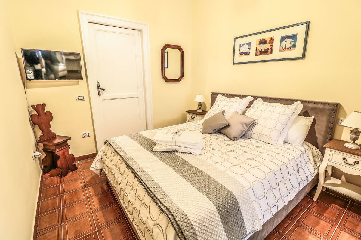 Una cama bien hecha con una colcha estampada, una mesita de noche con lámpara y un televisor montado en la pared.