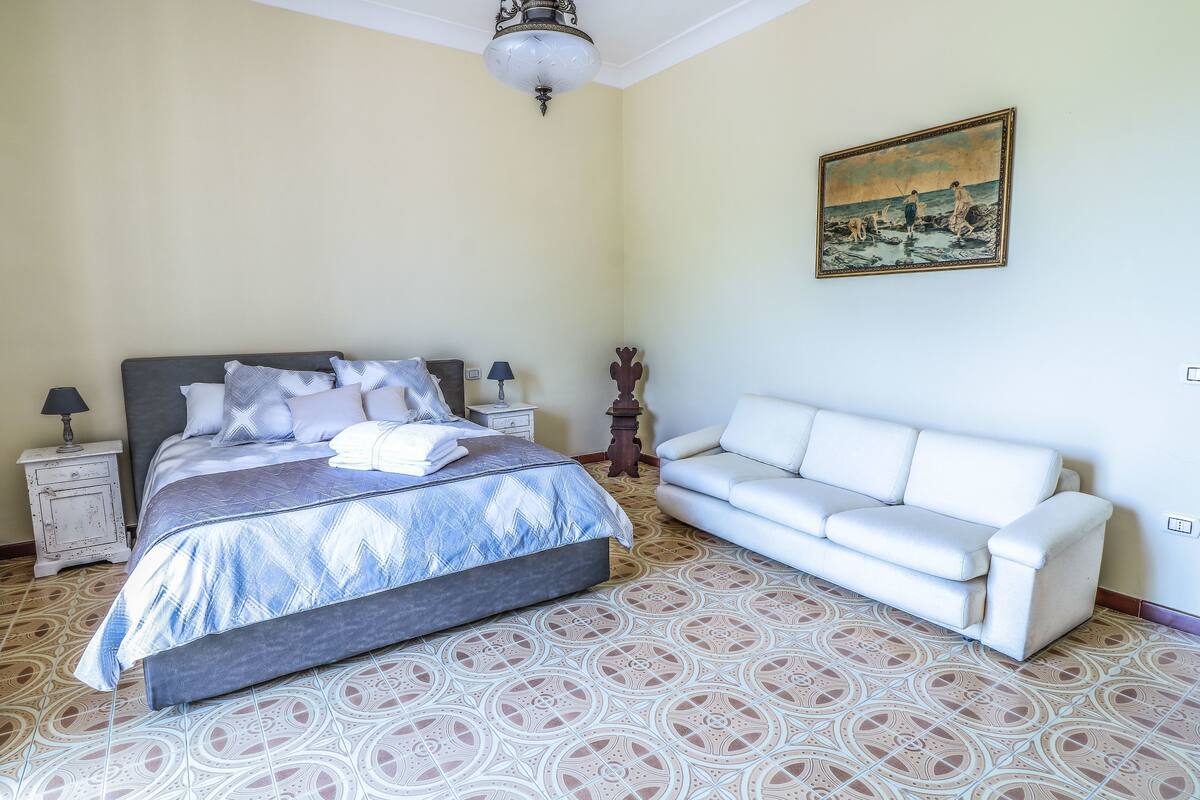 Un dormitorio con cama, un sofá blanco, baldosas de suelo estampadas, un cuadro en la pared y una araña.
