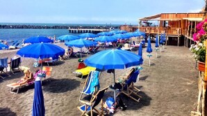A beach with numerous blue umbrellas and lounge chairs, a wooden building with a balcony, and a pier in the background. - Scognamiglio Suites (Ercolano)
