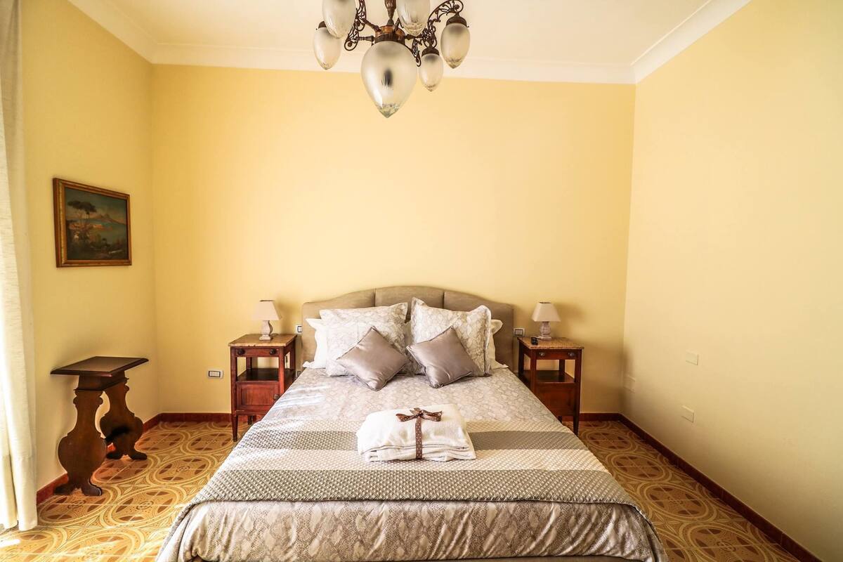 Una cama bien hecha con una colcha estampada y almohadas, dos mesitas de noche con lámparas, una araña y un cuadro en la pared.