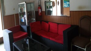 Een kamer met een rode bank, een zwarte salontafel en een rieten stoel. Een brandblusser hangt aan de muur en boven de bank is een spiegel.