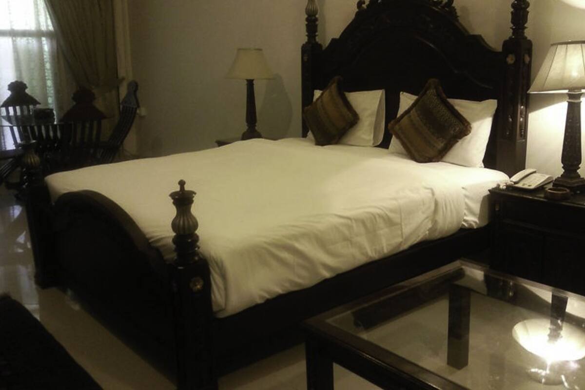 A neatly made bed with a wooden headboard, a bedside table with a lamp, and a glass coffee table in a hotel room.