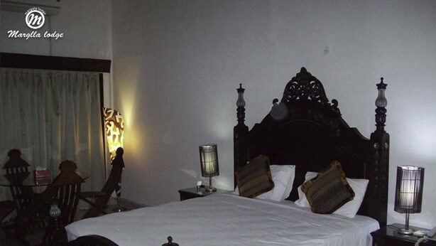 A hotel room with a large bed, two bedside tables, a chair, a lamp, and a wall-mounted light fixture. - Margalla Lodge (Islamabad)
