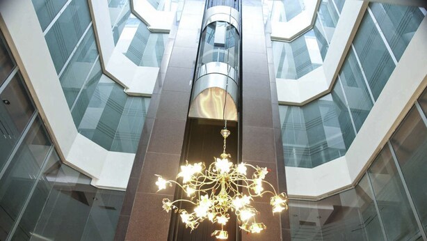 A modern interior with a central elevator shaft, glass walls, and a decorative chandelier. - Hotel Pak Heritage (Lahore)