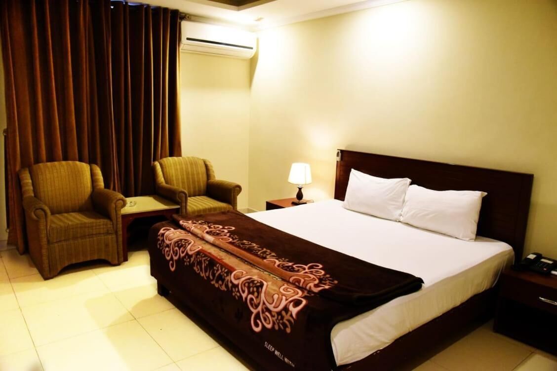 A hotel room with a bed, two armchairs, a small table, a nightstand with a lamp, and an air conditioning unit.