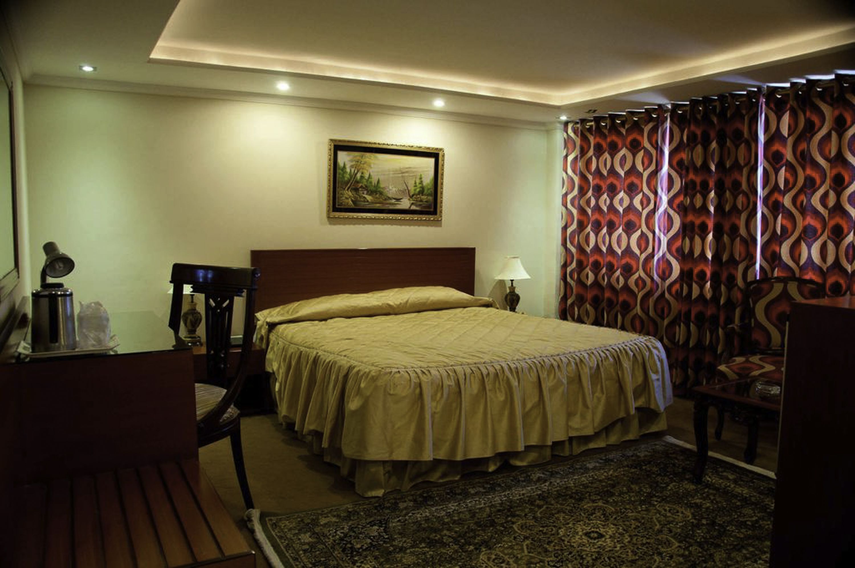 A hotel room with a large bed, a desk with a lamp, a chair, a nightstand, and a painting on the wall.