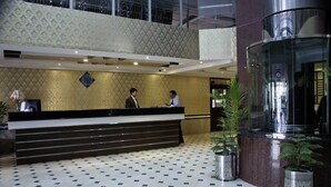 A hotel reception area with a check-in counter, two receptionists, a patterned wall, a glass elevator, and potted plants. - Hotel Pak Heritage (Lahore)