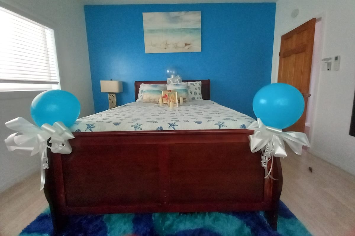 A bedroom with a wooden bed, blue walls, a beach-themed bedspread, and two blue balloons.