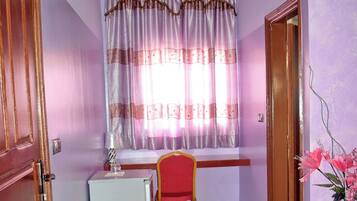 A room with a wooden ceiling, a window with curtains, a small white refrigerator, a red chair, and a door leading to another room.
