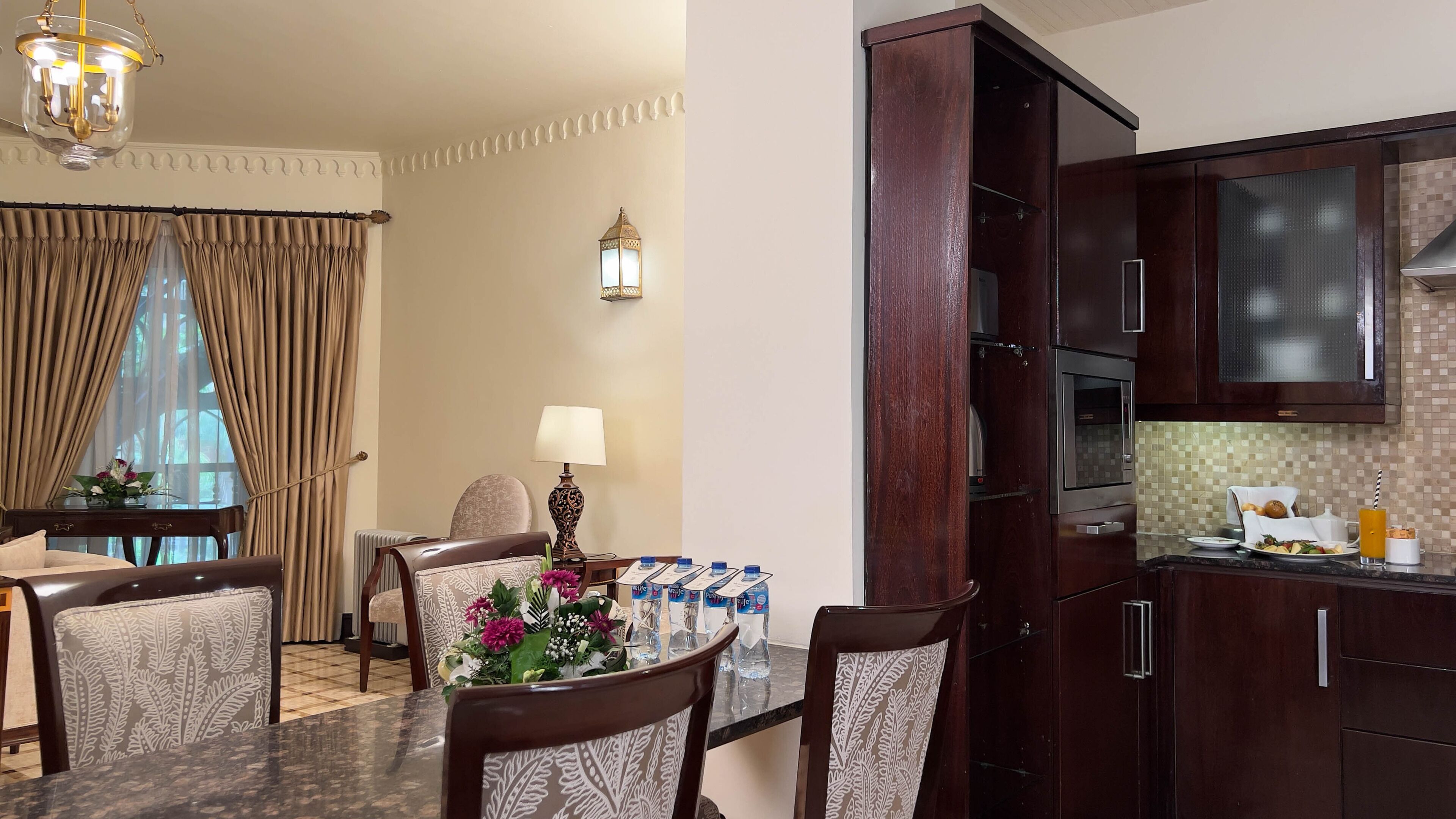 Habitación de hotel con comedor, cocina con armarios de madera oscura, mesa con sillas y una lámpara decorativa.