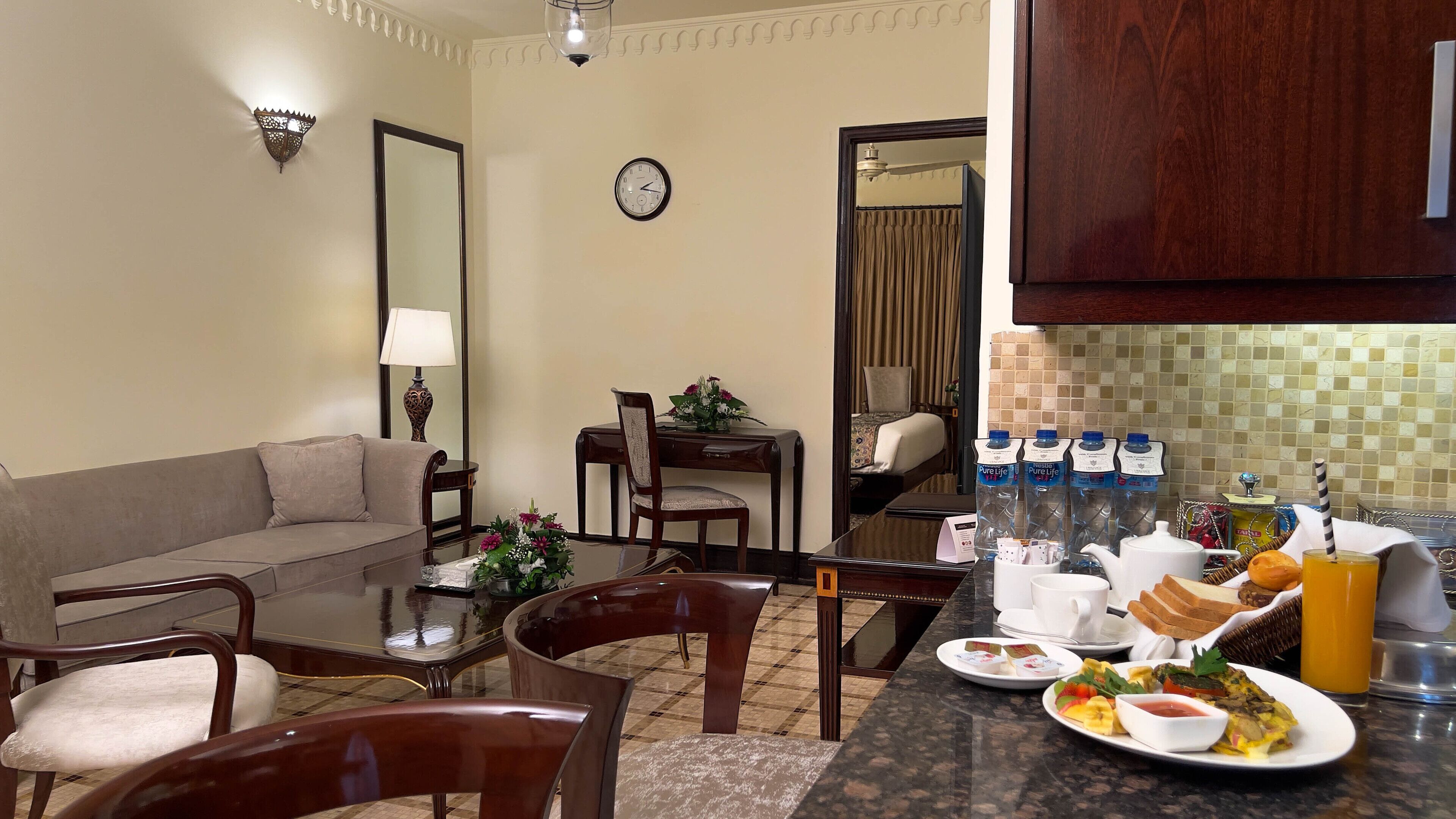 Habitación de hotel con comedor, sofá, mesa con alimentos para el desayuno y un reloj en la pared.