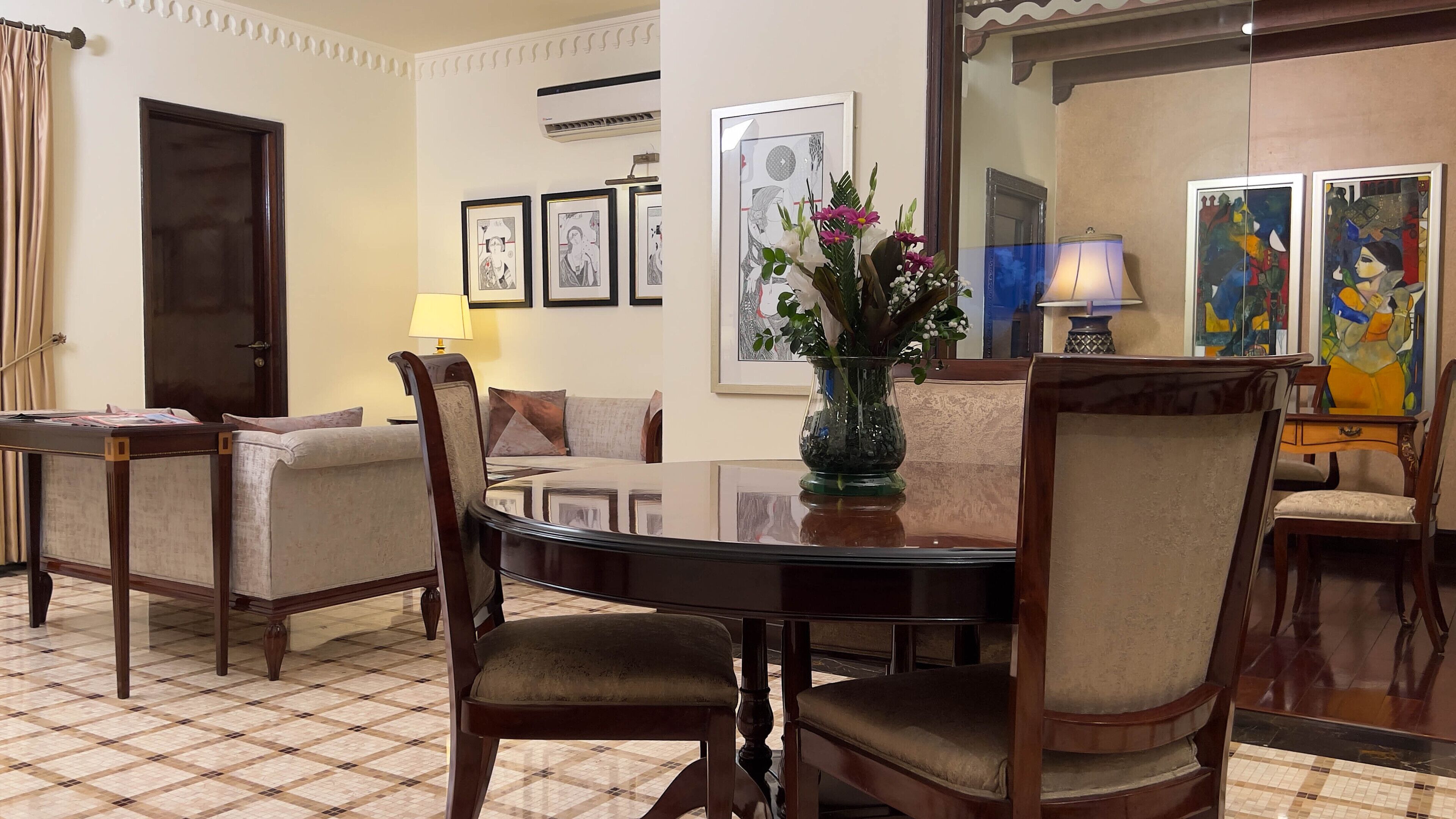 A dining area with a round table, chairs, a vase with flowers, framed artwork on the wall, and a door in the background.
