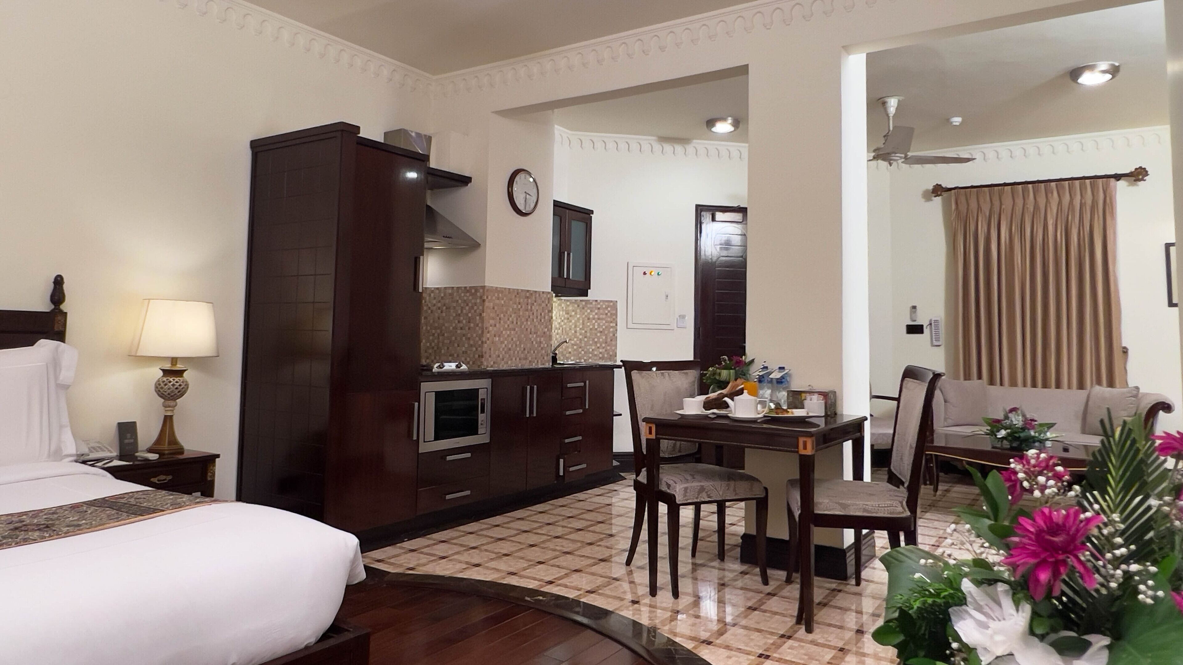 A hotel room with a bed, a desk with chairs, a kitchenette, and a living area with a sofa and curtains.