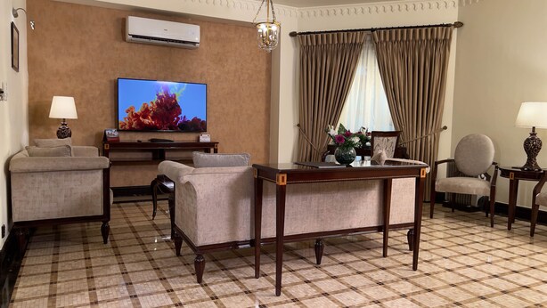 Habitación de hotel con televisor de pantalla plana, un escritorio de madera, dos sillones, una mesa de centro y un amplio ventanal con cortinas.