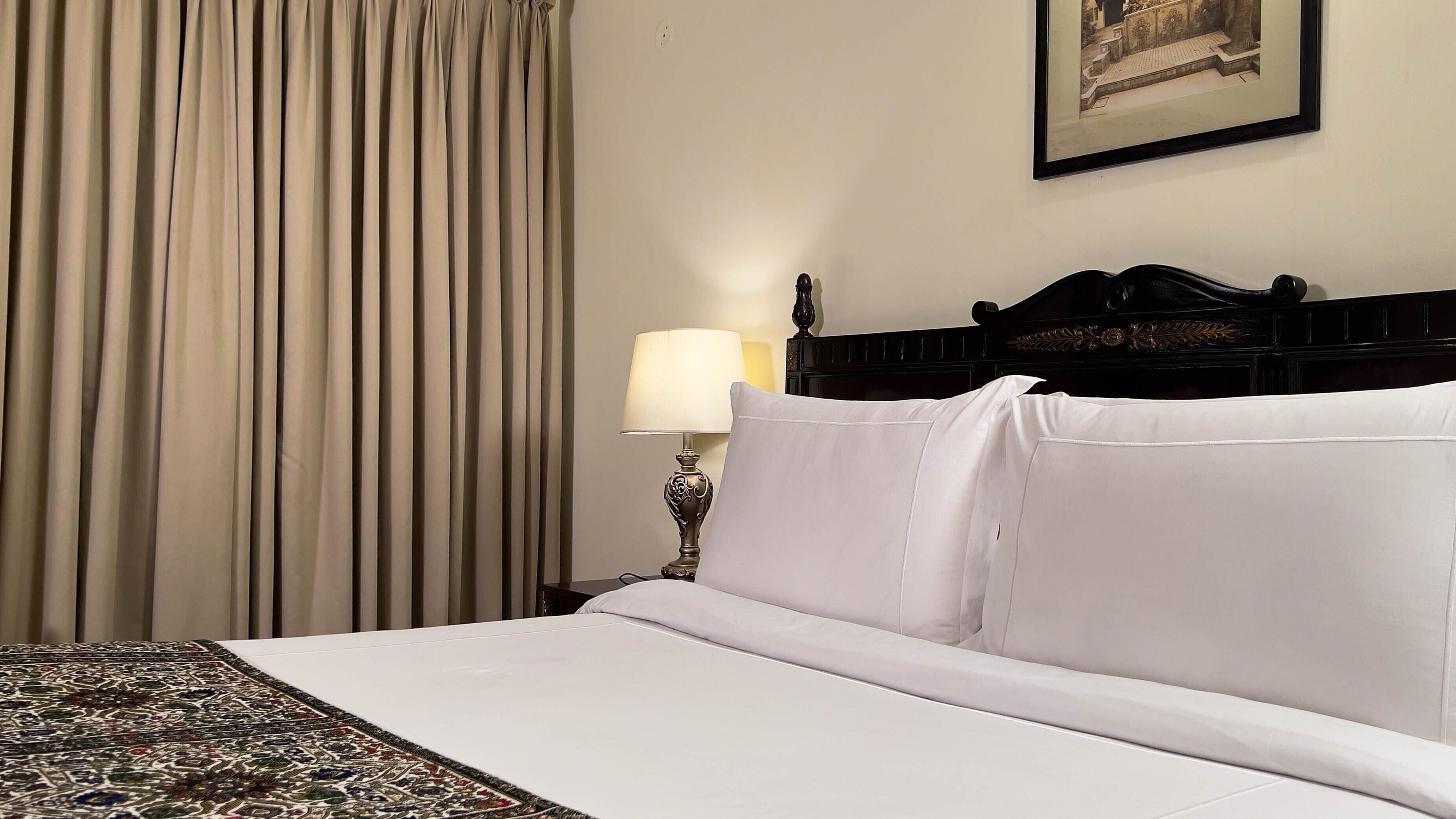A neatly made bed with white linens, a patterned rug, a lamp, and a framed picture on the wall.