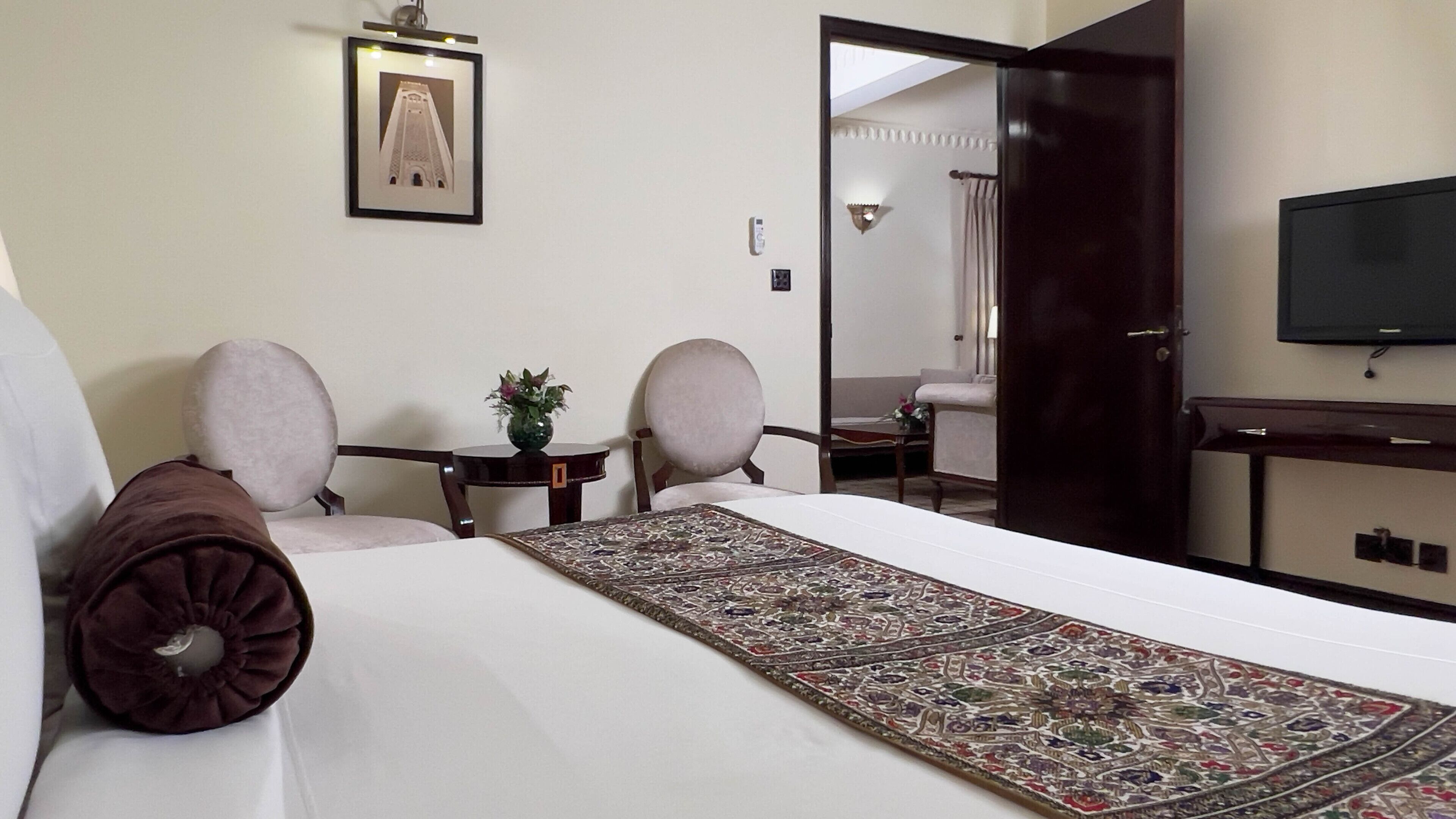 Ein Hotelzimmer mit einem großen Bett, zwei Sesseln, einem kleinen Tisch mit einer Vase, einem gerahmten Bild an der Wand, einem Fernseher und einer Tür zu einem anderen Raum.