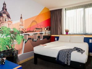 A hotel room with a bed, a desk, a chair, a window with curtains, a wall mural, and a potted plant. - Ibis Styles Budapest Citywest (Budapest)