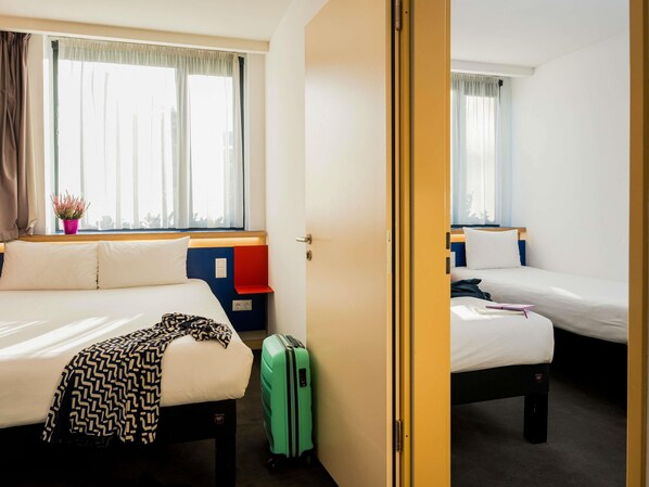 A hotel room with two beds, a window with curtains, a small plant, and a suitcase.