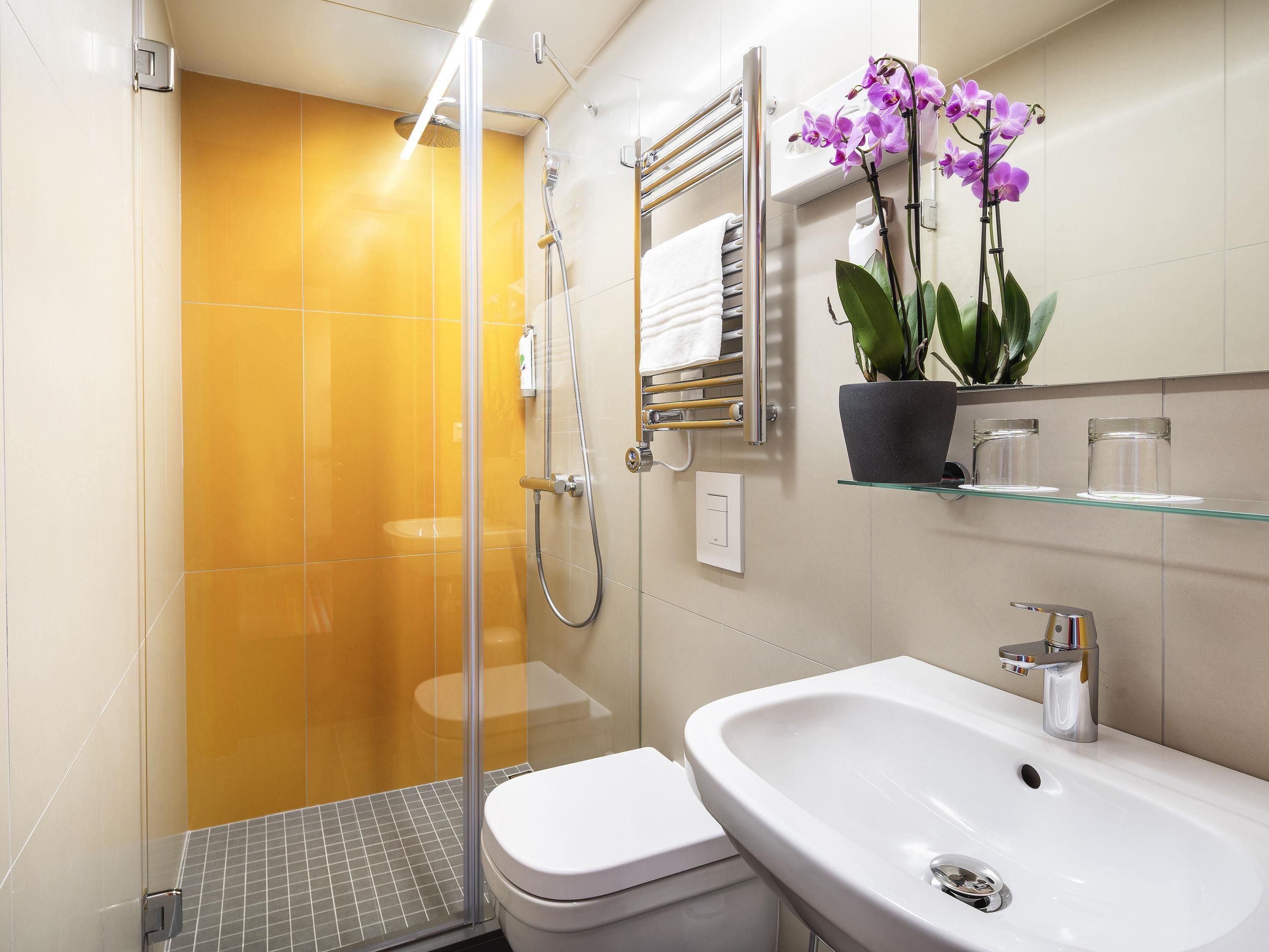 Une salle de bain avec une douche à parois de verre, des toilettes blanches, un lavabo avec un robinet chromé et une orchidée en pot posée sur une étagère.