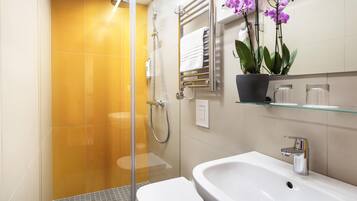 Une salle de bain avec une douche à parois de verre, des toilettes blanches, un lavabo avec un robinet chromé et une orchidée en pot posée sur une étagère.