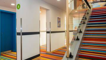 Un pasillo de hotel con una alfombra colorida a rayas, una puerta azul y una escalera con barandilla de vidrio.