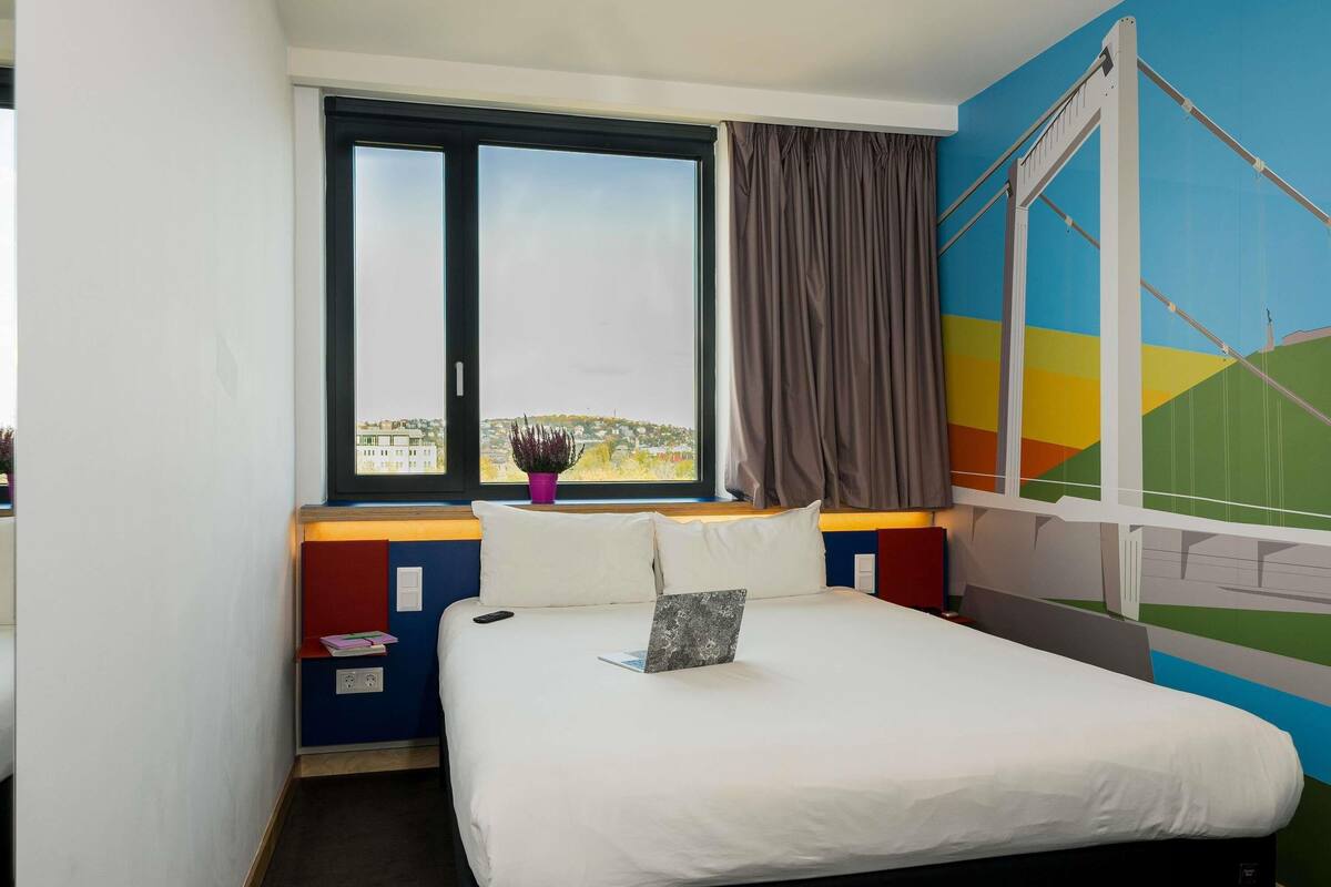 A hotel room with a bed, a window with curtains, a view of a cityscape, and a colorful wall.