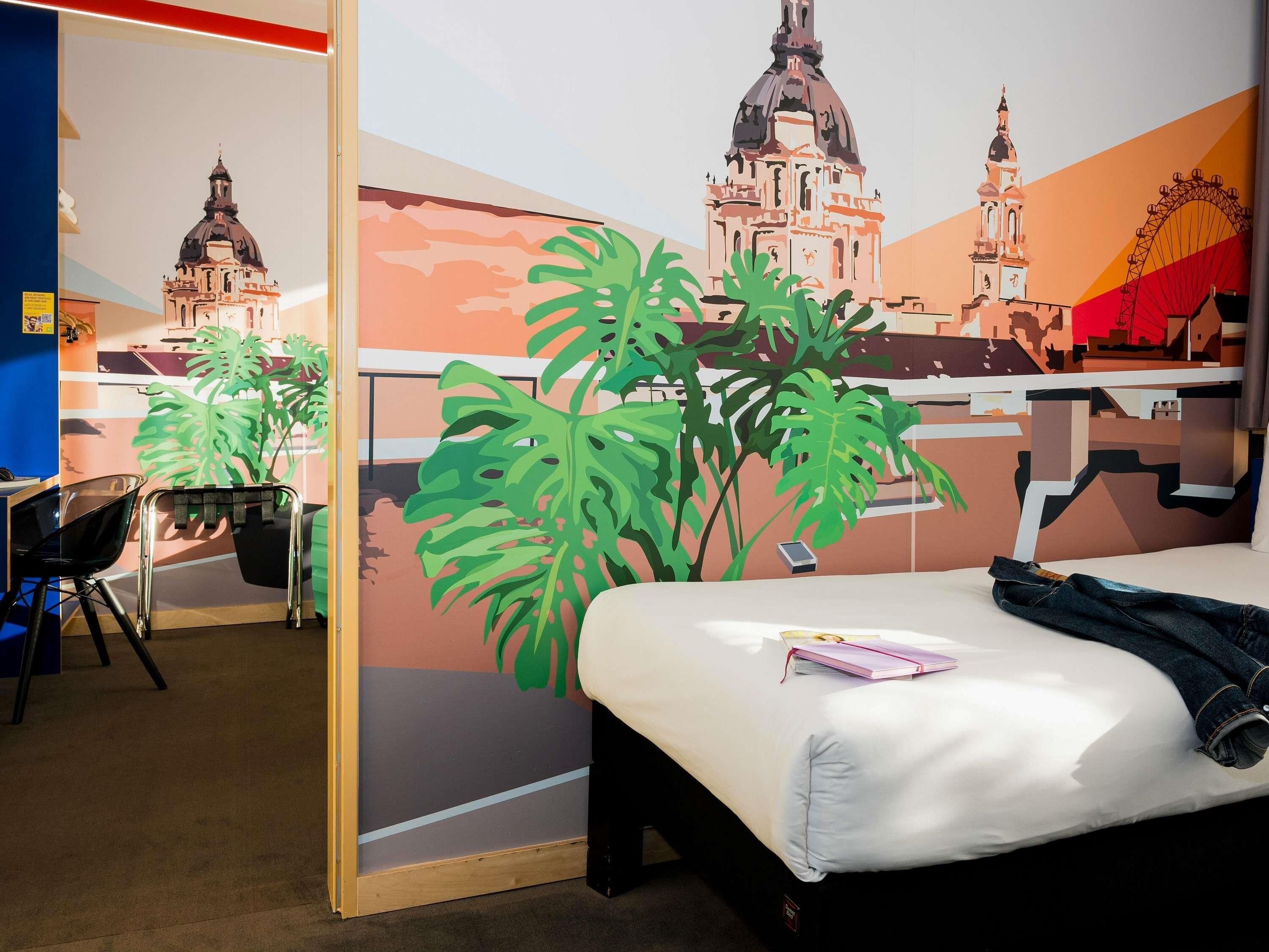 A hotel room with a bed, a desk, and a chair. There are large wall murals depicting buildings and a Ferris wheel.