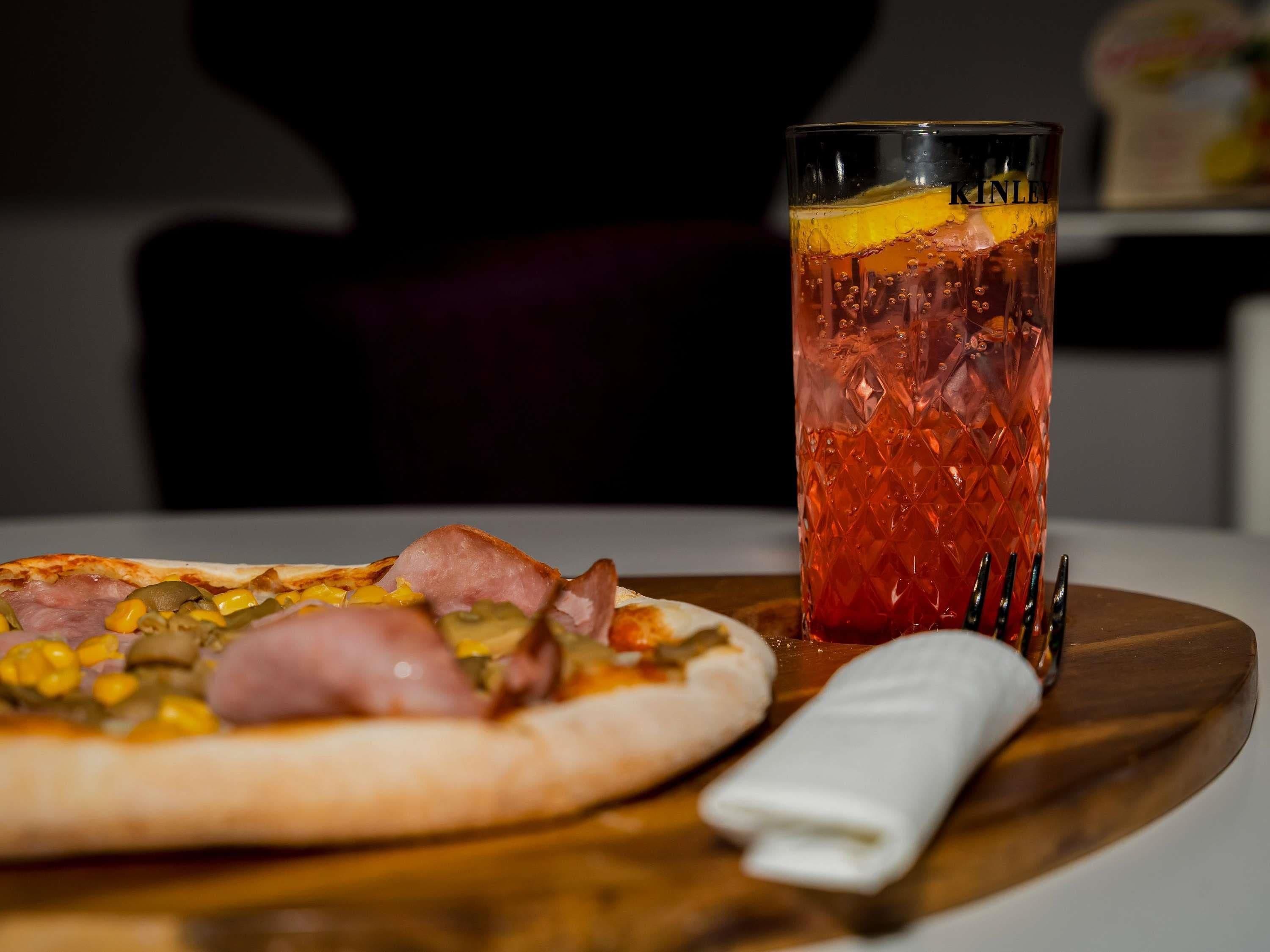 Una pizza con jamón y verduras sobre una tabla de madera, un vaso alto con una bebida roja y un tenedor sobre un mantel blanco.