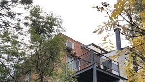 A modern multi-story building with a balcony on the upper floor, surrounded by greenery.