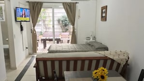 A bedroom with a bed, a TV, a window with curtains, and a dining area with a table and chairs.