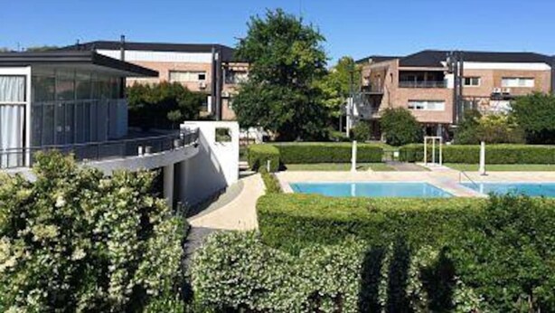 A modern residential building with a swimming pool, surrounded by well-maintained hedges and a clear blue sky.
