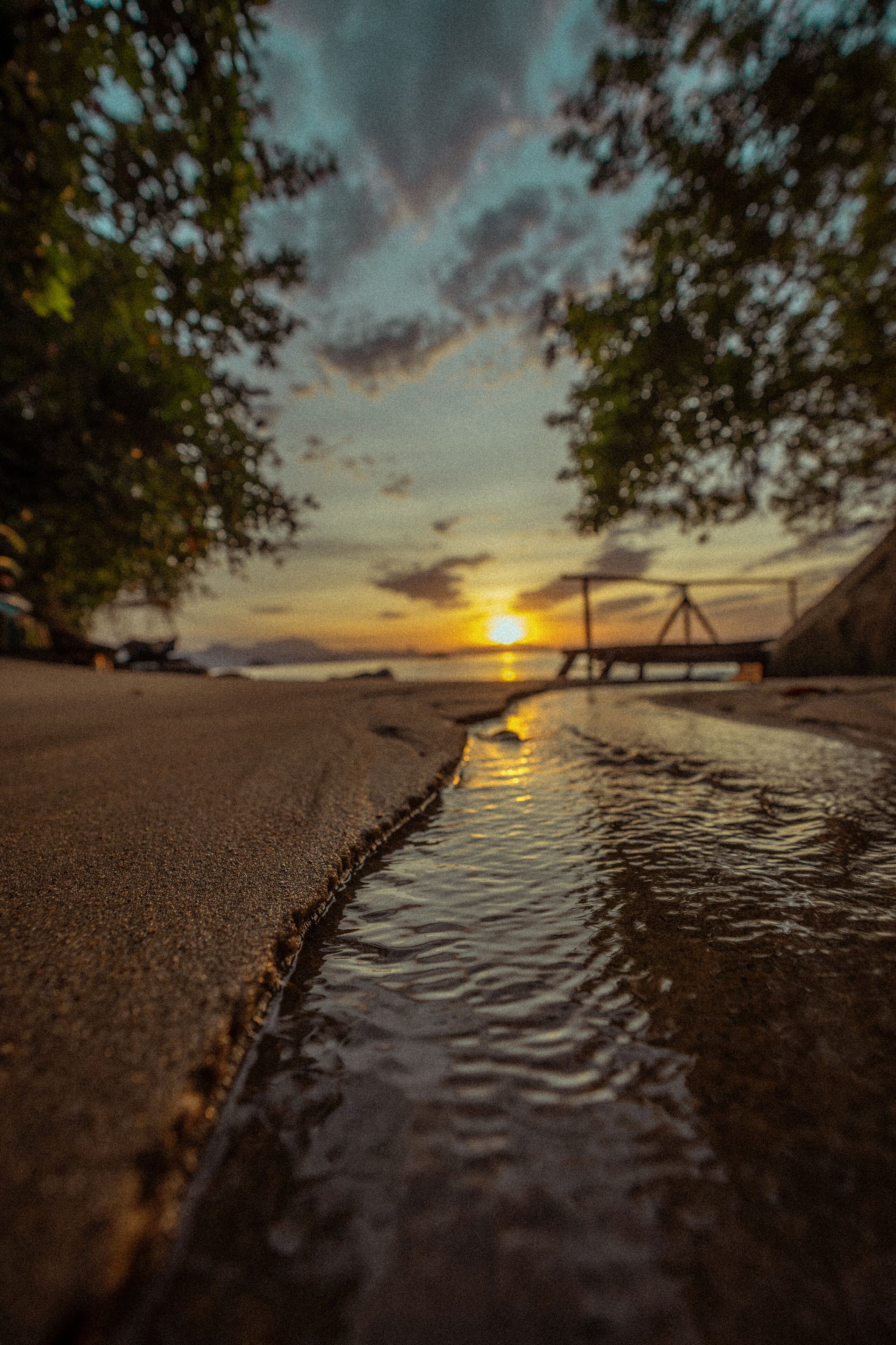 Escena de playa con un cuerpo de agua tranquilo, un puente de madera y un atardecer al fondo.
