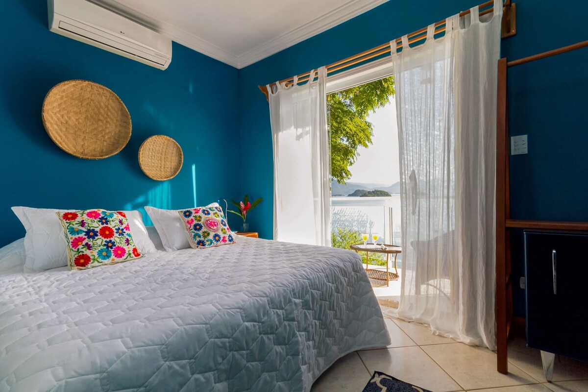 Un dormitorio con cama, almohadas, una mesita de noche, lámpara, ventana con cortinas y vista al exterior.