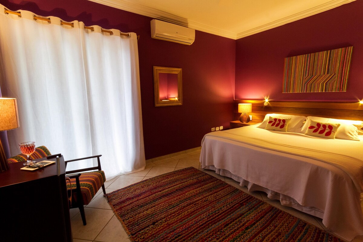 Habitación de hotel con cama, escritorio, silla, ventana con cortinas, aire acondicionado de pared y un cuadro.