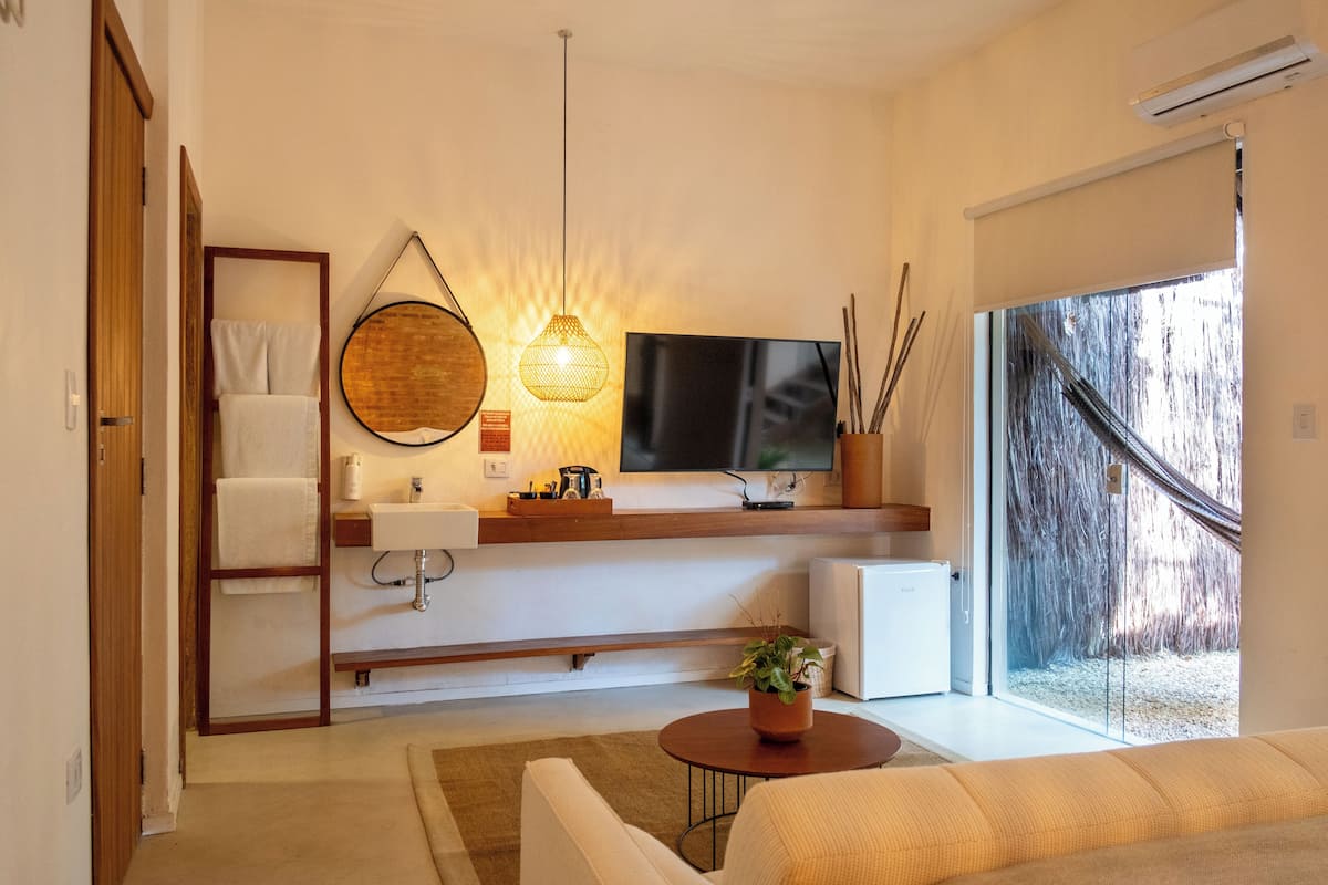 Un salón moderno con televisor de pantalla plana, una lámpara redonda fijada a la pared, una mesita con una planta y un amplio ventanal con vistas a árboles.