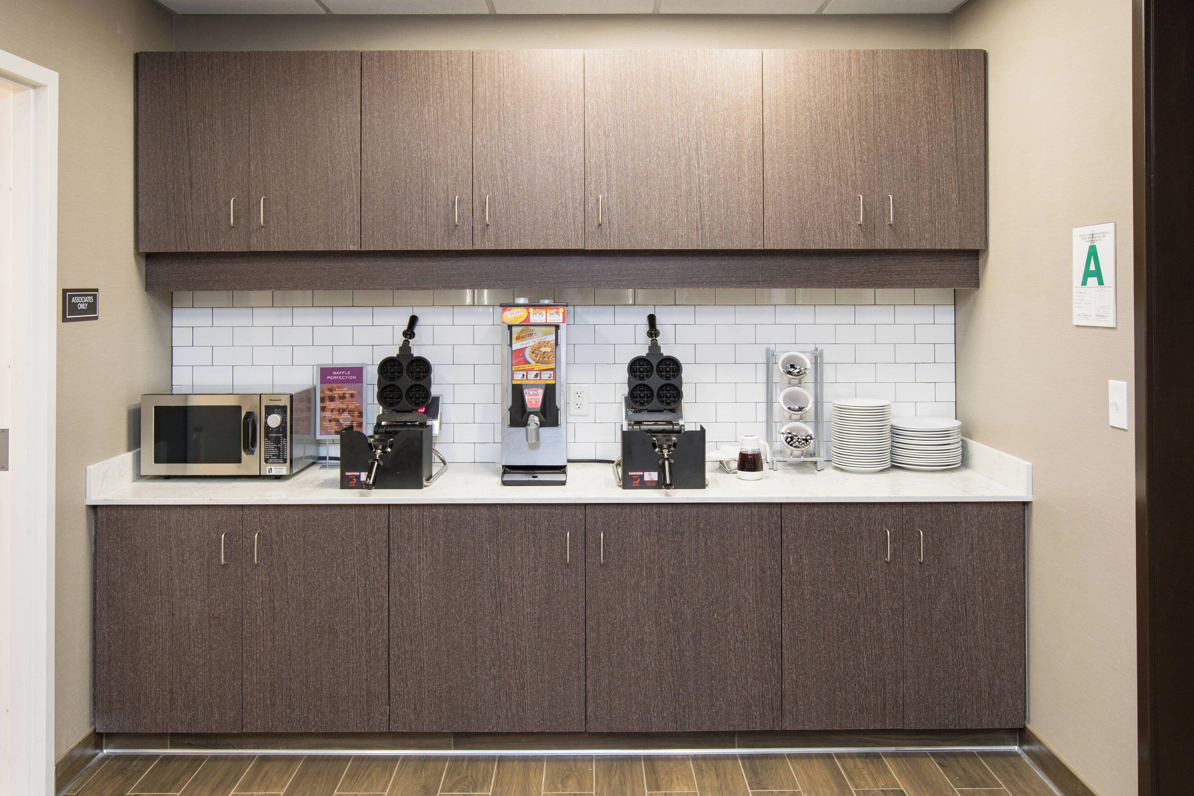 A modern kitchen with dark wood cabinets, a white tiled backsplash, and various appliances including a microwave, coffee maker, and waffle maker.