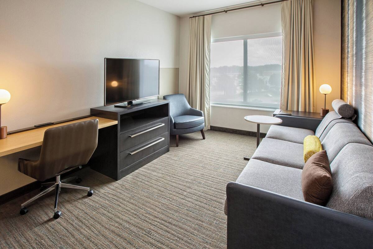 Ein Hotelzimmer mit einer Sitzecke, einem Schreibtisch mit Fernseher, einem Sessel, einem kleinen Tisch und einem Fenster mit Vorhängen.