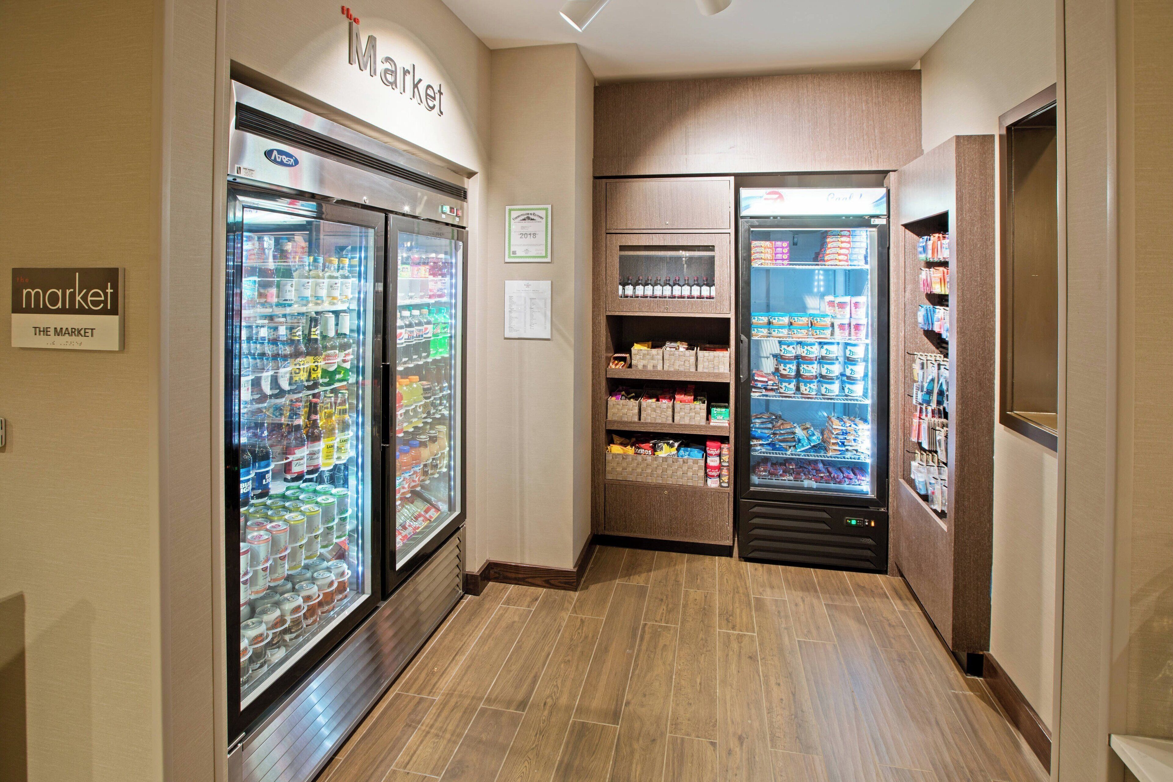 Un corridoio con vetrine refrigerate piene di bevande e snack, pavimento in legno e un cartello che indica una zona dedicata al mercato.