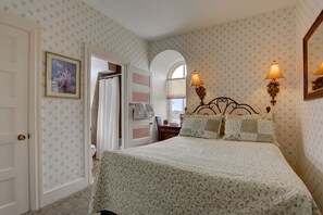 A bedroom with a floral patterned bedspread, a white door, a framed picture on the wall, and two wall-mounted lamps.