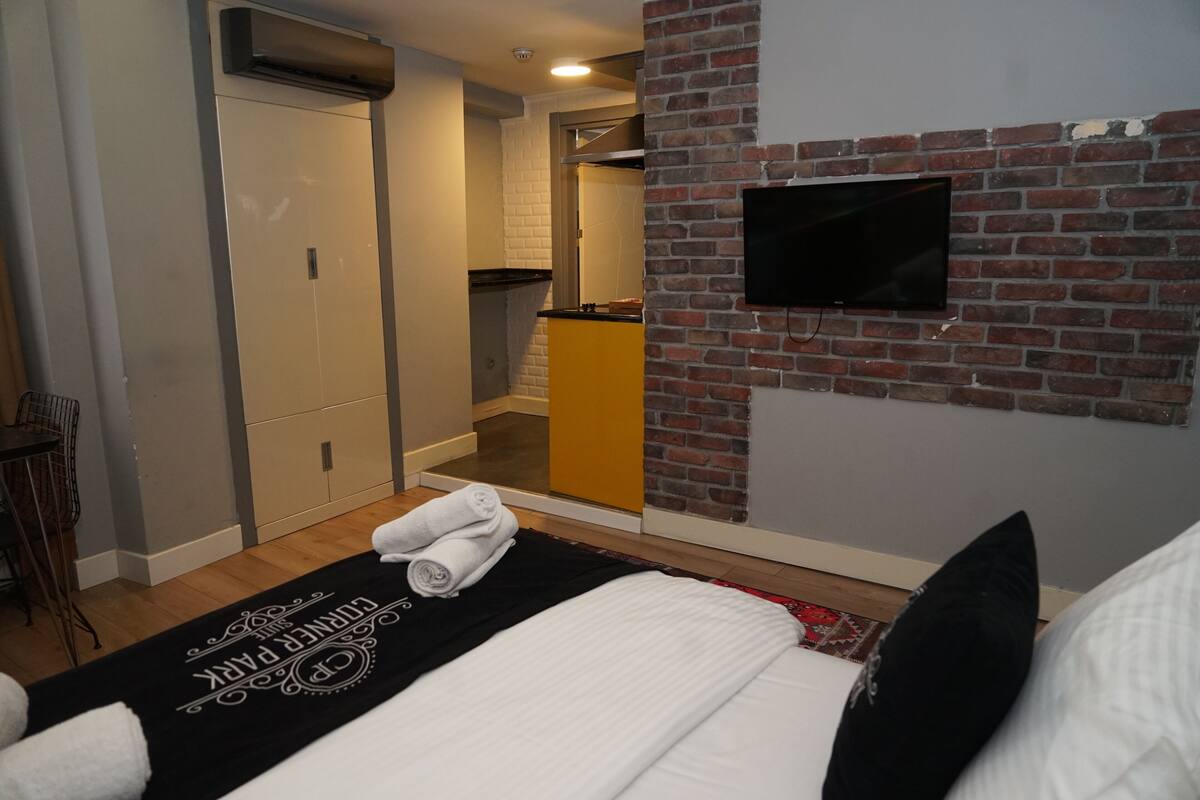 A hotel room with a bed, a TV mounted on a brick wall, a bathroom with a yellow cabinet, and a door leading to another room.