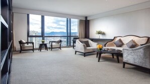 Amplia habitación de hotel con un ventanal grande que ofrece vistas a la ciudad, un sofá cómodo, una mesita de centro y una zona de estar con sillas.