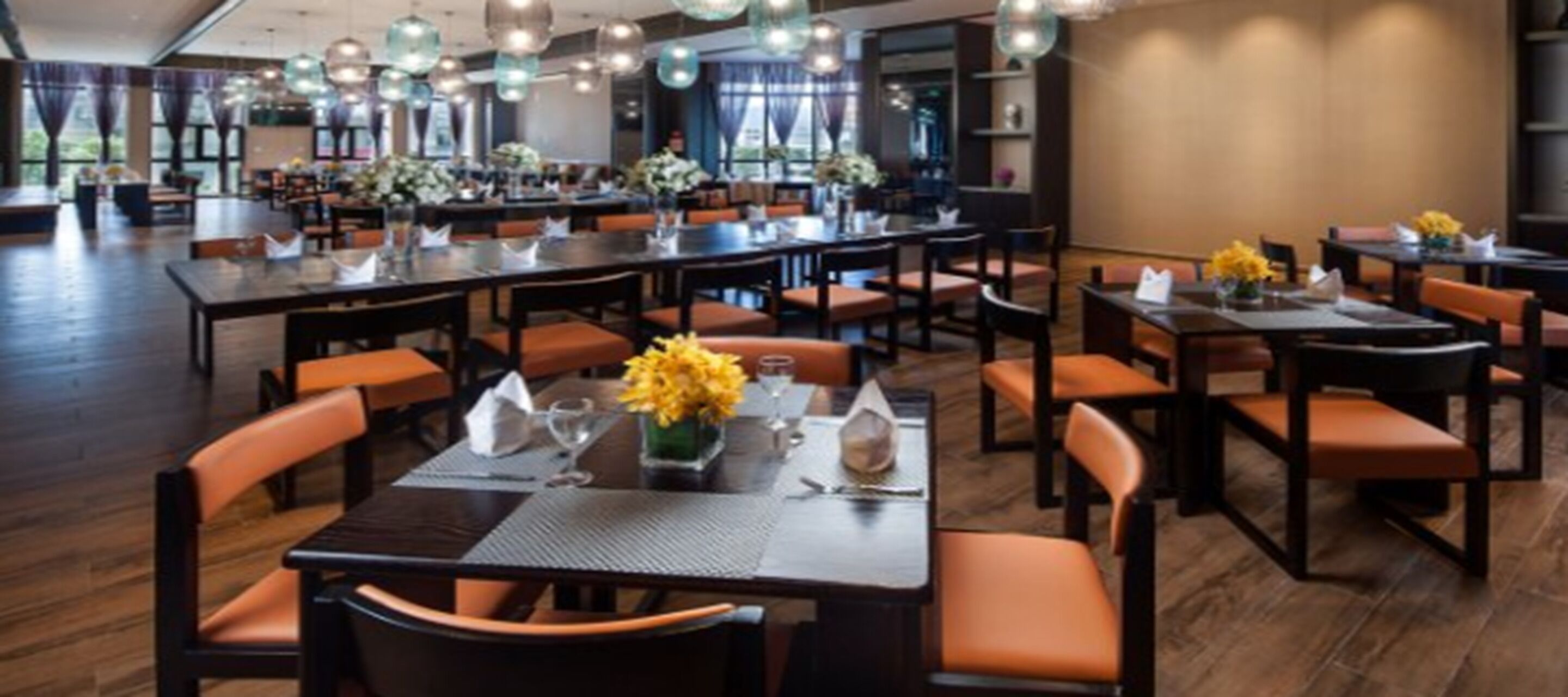 A restaurant with multiple tables and chairs, arranged in a spacious layout with a high ceiling and pendant lighting.