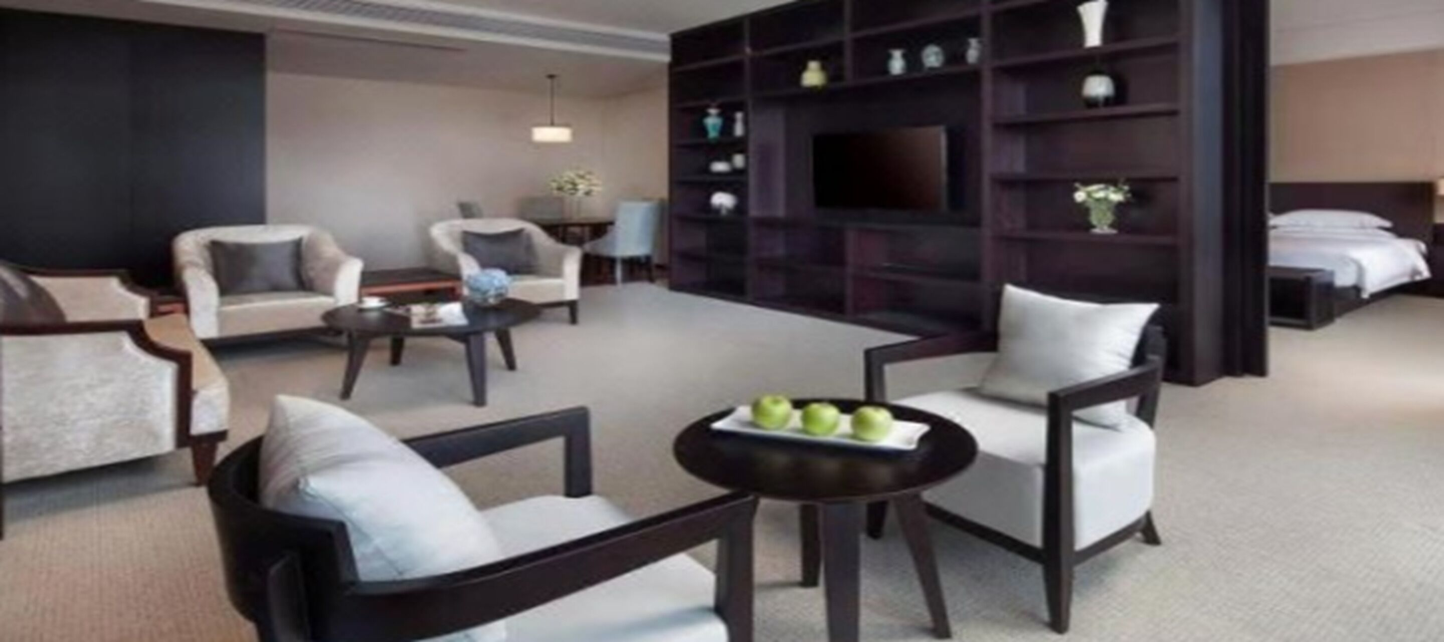A modern hotel room with a living area featuring a sofa set, a coffee table with apples, and a dark wooden bookshelf with decorative items.