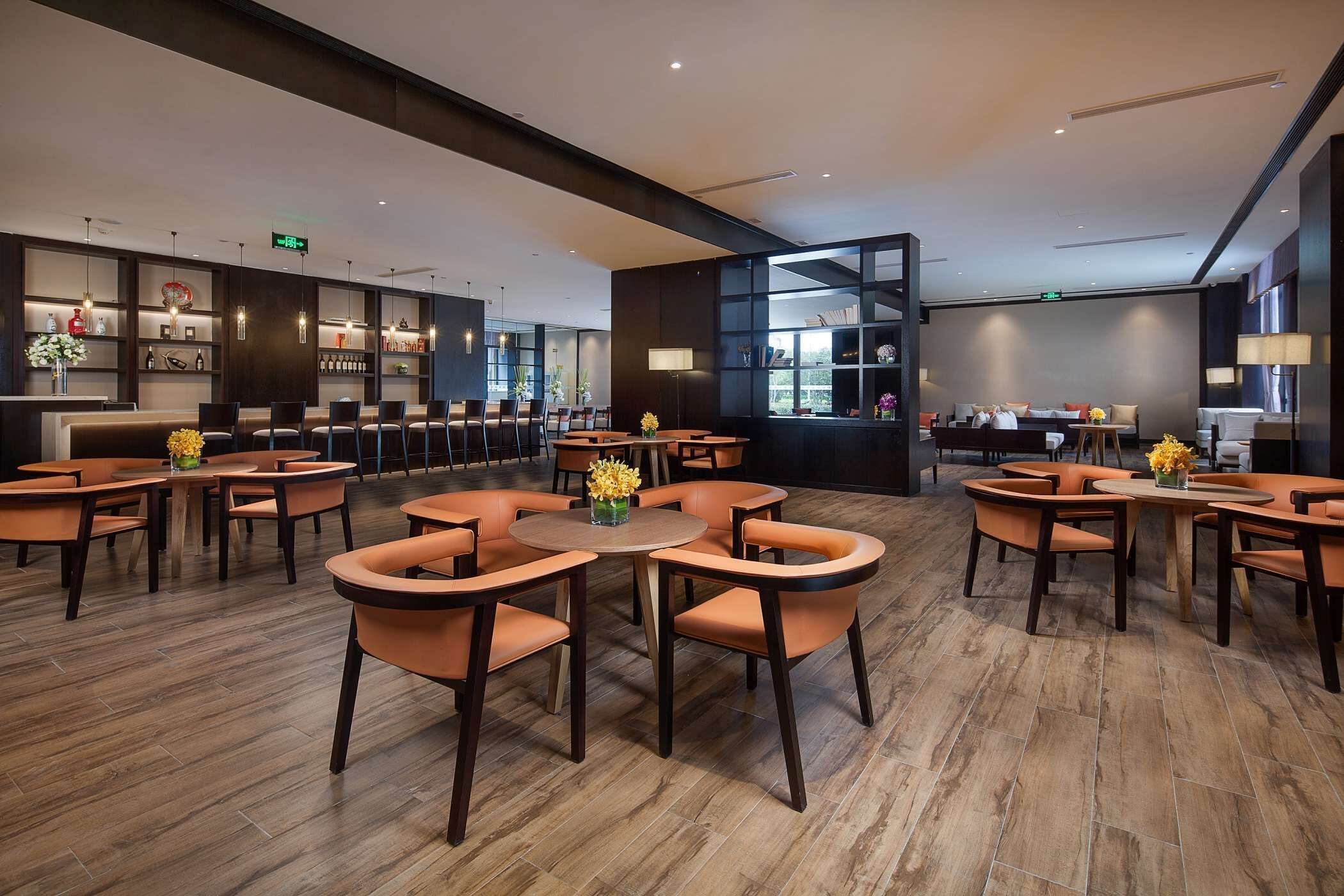 A modern hotel lobby with round tables and orange chairs, a reception desk, and a bookshelf.