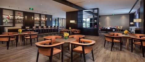 A modern hotel lobby with round tables and orange chairs, a reception desk, and a bookshelf.