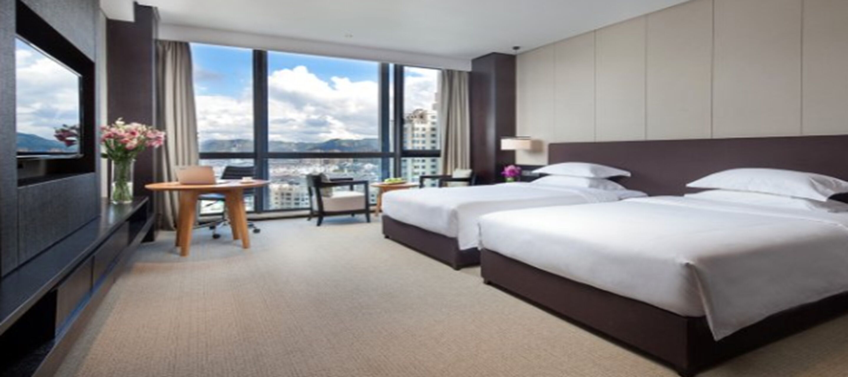 A hotel room with two beds, a desk, a chair, a TV, and a large window offering a city view.