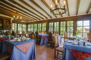 A dining area with tables set for service, wooden beams on the ceiling, and large windows with a view of greenery outside.