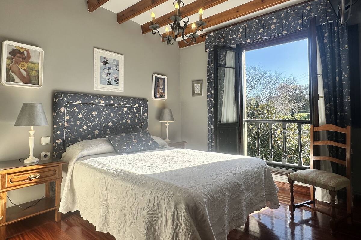 A bedroom with a large bed, a nightstand, a chair, and a window with a view of trees.