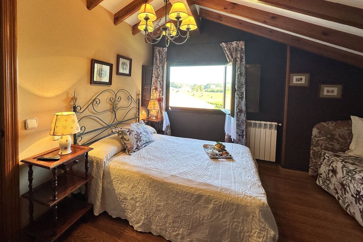 A bedroom with a large bed, a bedside table with a lamp, a window with curtains, and a radiator.